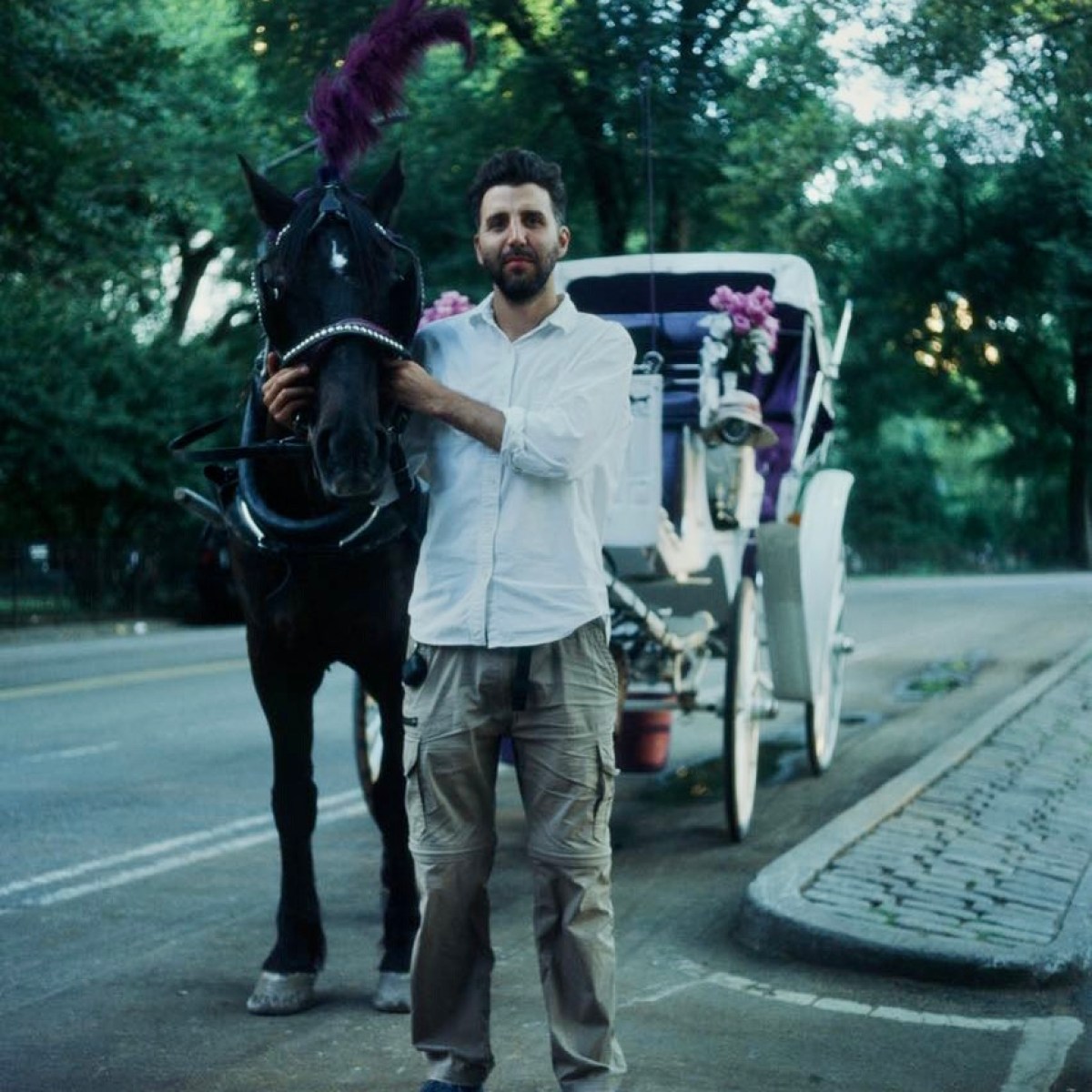 Man in white shirt stands next to a horse with carriage on a tree-lined street.