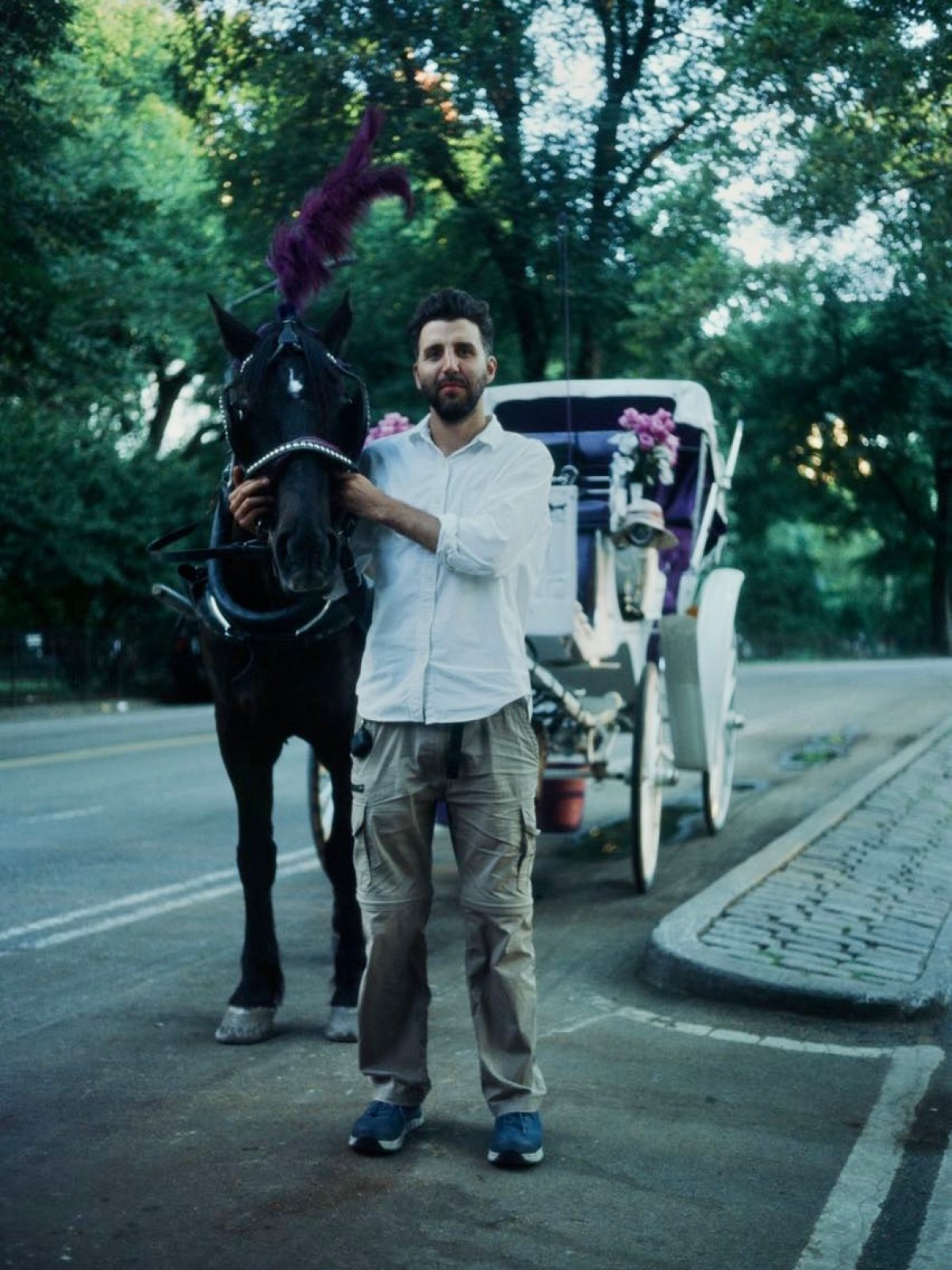 Man in white shirt stands next to a horse with carriage on a tree-lined street.