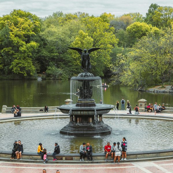 People around a large fountain in a park with trees and a pond.